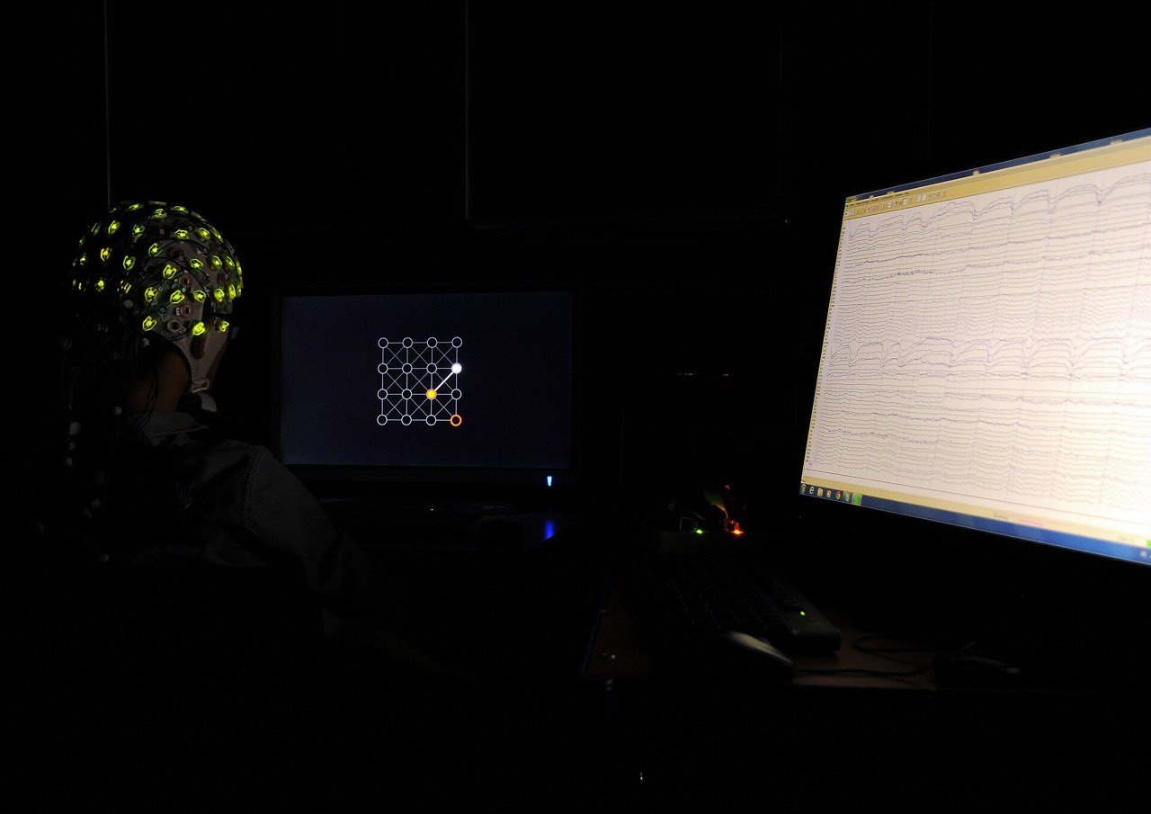 Participante de experimento com touca de EEG diante de uma tela, com a atividade cerebral exibida em outro monitor